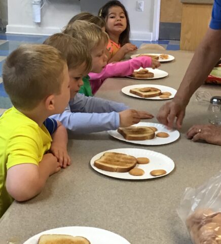 Children sitting at a table with plates of toasted sandwiches and cookies, being served by an adult.