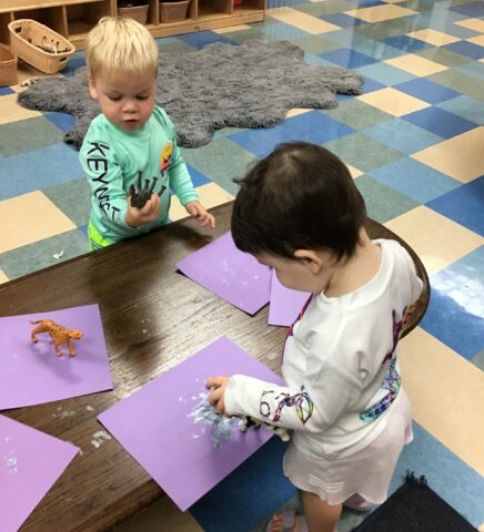 Two toddlers engaged in a craft activity with purple paper and paint in a playroom. One holds a toy dinosaur.