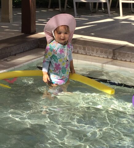 A child in a sun hat and floral swimsuit stands in a shallow pool, holding a yellow pool noodle.