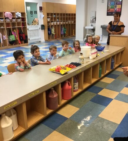 Children sitting at a table with snacks and ingredients in a classroom. A teacher stands nearby, and backpacks and cubbies are visible in the background.