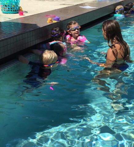 Four children wearing goggles are in a swimming pool, holding onto the edge. An adult in the water is facing them, likely giving a swimming lesson. A basket and toys are on the poolside.