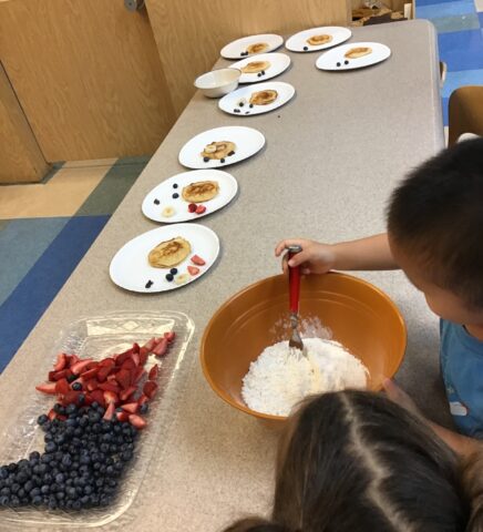 Two children mix batter in a bowl. On the table, plates with pancakes, scattered blueberries, and strawberries are displayed. A tray with more berries is nearby.