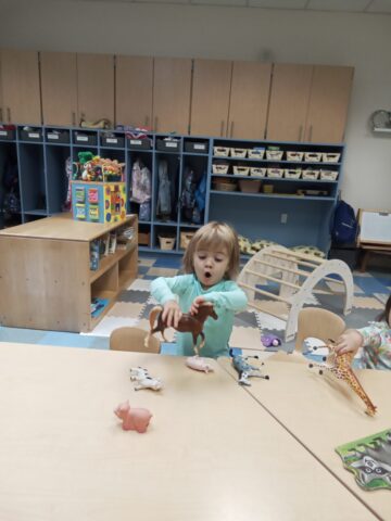 A child plays with animal toys at a classroom table. Shelves with bins and backpacks are visible in the background.