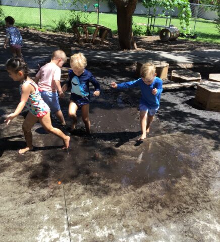 Children playing and splashing in a muddy puddle on a sunny day in a fenced outdoor area.