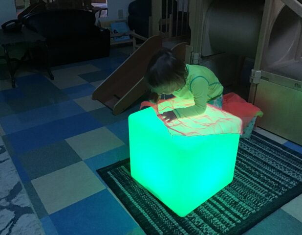 A child in pajamas kneels on a glowing green cube in a dimly lit room with a checkered floor.