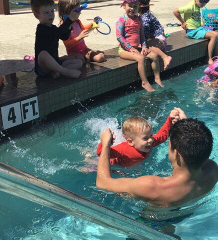 Children are sitting by the pool, watching one child being lifted and splashed in the water by an adult. The pool depth is labeled 4 FT.