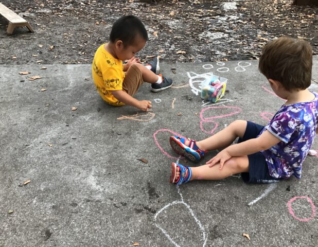 Two children sit on a concrete surface drawing with colorful chalk. A box of chalk is nearby, and various doodles cover the ground.