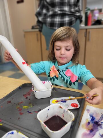 A child in a floral shirt paints a cardboard tube with a brush, surrounded by paint containers on a table.