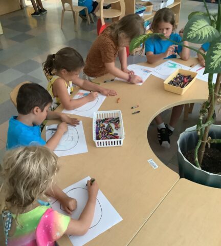 Five children sit around a table drawing with crayons on paper in a classroom.