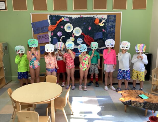 A group of children in colorful outfits stand in a line, each holding paper plate masks decorated with drawings. They are in a classroom with crafts displayed on the wall behind them.