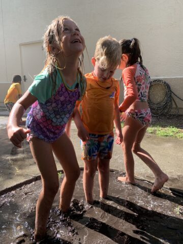 Children play and splash in a muddy puddle, wearing swimsuits, with sunlight casting shadows on the ground.