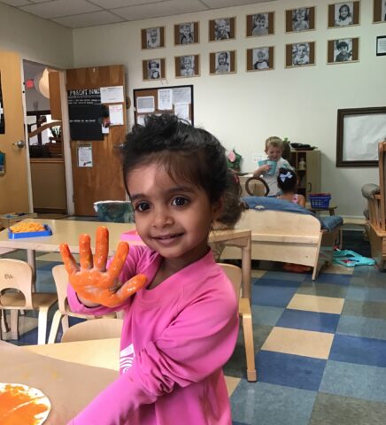 A young child in a pink shirt shows a paint-covered hand, smiling, in a classroom with pictures on the wall. Another child can be seen playing in the background.