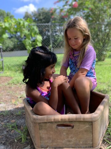 Two children sit playfully in a wooden box outdoors, surrounded by greenery and a chain-link fence.