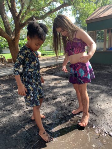 Two children in colorful outfits stand barefoot in a muddy puddle outdoors, with trees and a building in the background.