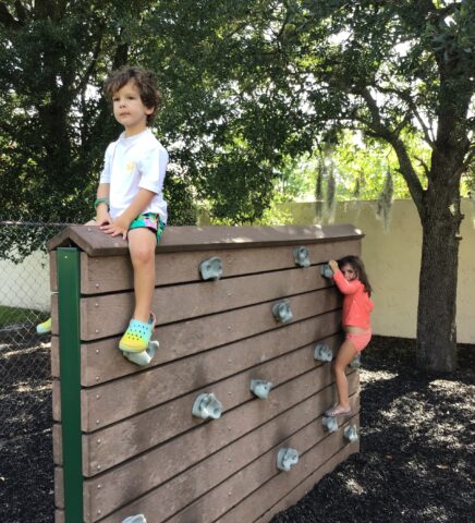 Two children climbing on a wooden climbing wall in a playground. One sits on top while the other hangs on the side. Trees and a chain-link fence are in the background.