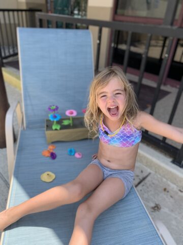 A child in a colorful swimsuit smiles while sitting on a lounge chair, with toy flowers placed beside them.