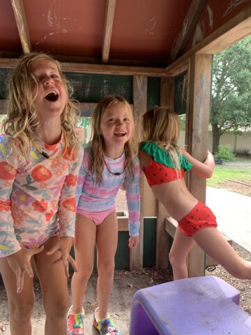 Three young children playing in a wooden playhouse, wearing colorful swimsuits and smiling.