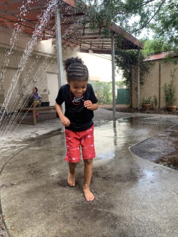 A child in red shorts and a black shirt plays in water sprays outdoors. Another person sits on a bench in the background. Trees and a covered area surround them.