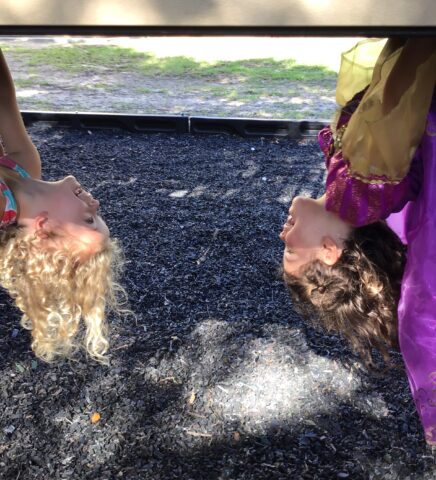 Two children hang upside down on playground equipment, smiling at each other. One wears a colorful dress, and the other wears a purple outfit.