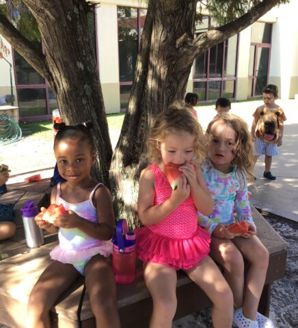 Three children sitting on a bench under a tree, eating watermelon slices. They are dressed in colorful summer outfits.