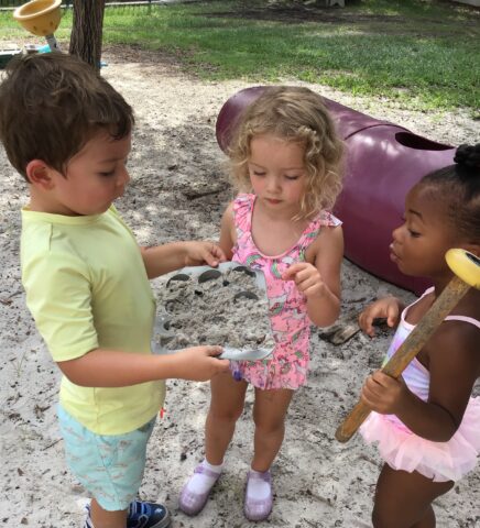 Three children playing outdoors with sand in a muffin tin and toy tools. A playground tunnel is in the background.