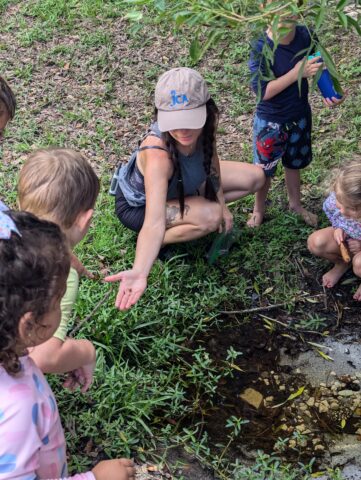 A woman shows something in her hand to a group of children gathered around a small water puddle in a grassy area. They appear to be outdoors, wearing casual summer clothing.