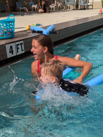 An adult and child are swimming in a pool using pool noodles. The water depth is marked as 4 feet. There's a basket and toys on the poolside in the background.