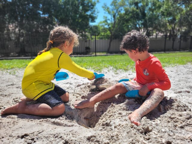 Two children play with blue shovels in a sandy area outside, surrounded by green grass and trees under a clear blue sky.