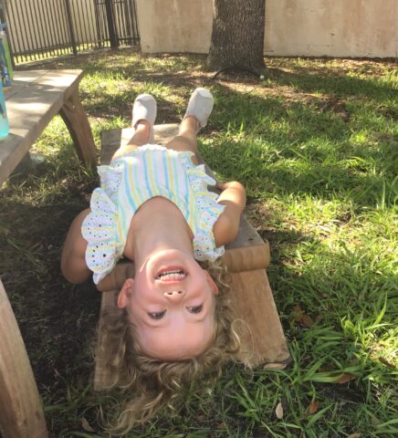 Child in a striped outfit lying upside down on a wooden bench, smiling widely with grass and a tree in the background.