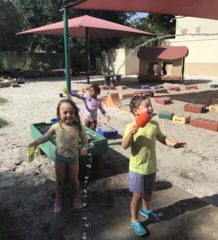 Three children play in a sandy playground with colorful blocks and water toys under shaded areas.