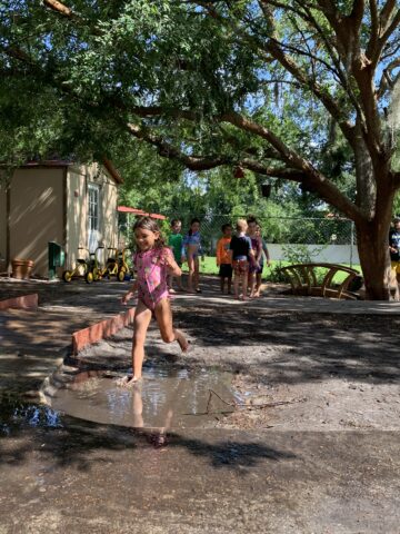 A young girl jumps over a muddy puddle in a sunny outdoor play area while several children watch in the background.