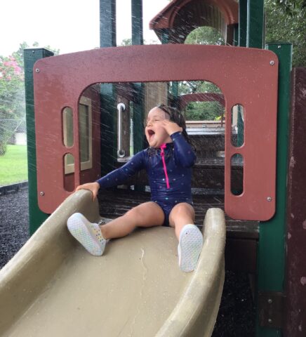 A child in a blue swimsuit sits laughing at the top of a playground slide while water sprays around them.