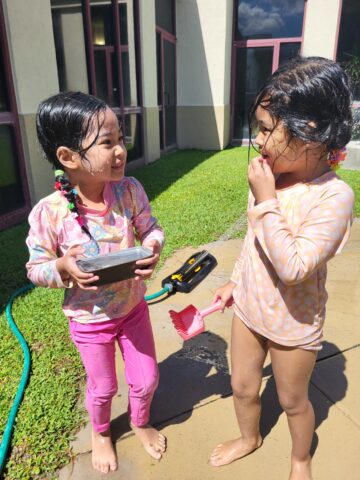 Two children with wet hair are playing outside near a building. One holds a rectangular object, and the other has a pink shovel. A green hose and a black tool are on the ground.