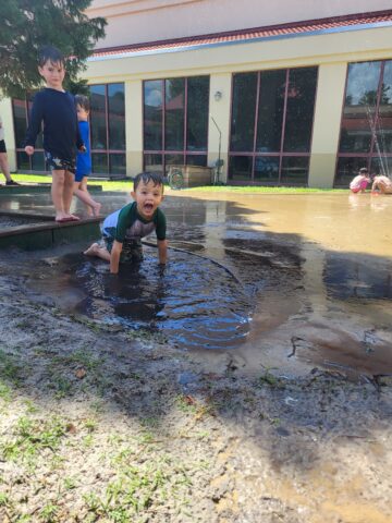 A child in a green shirt is playing in a muddy puddle, while another child stands nearby. A few more children can be seen in the background near a building with large windows.