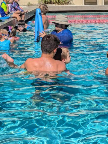 A swim instructor helps a young child in a pool, surrounded by other children and another adult. The atmosphere is lively and sunlit.