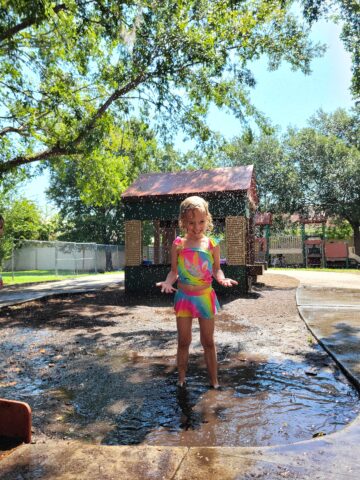 A child in a colorful swimsuit plays joyfully in a sunlit park, splashing water, with trees and a small building in the background.