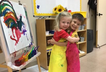 Two children hugging in front of an easel.