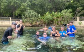 A group of people in swimwear standing and swimming in a clear, shallow pool surrounded by greenery. They are posing and giving thumbs-up gestures.