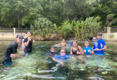 A group of people in swimwear standing and swimming in a clear, shallow pool surrounded by greenery. They are posing and giving thumbs-up gestures.