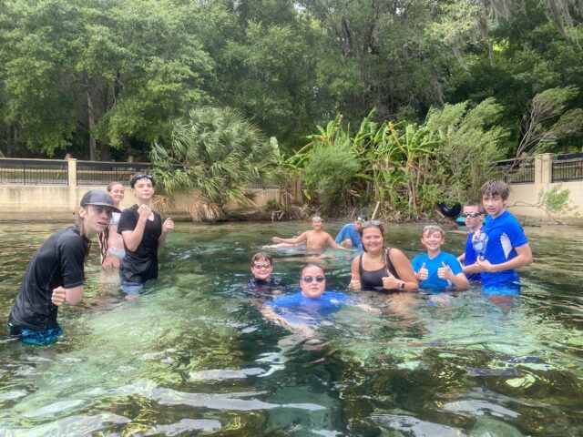 A group of people in swimwear standing and swimming in a clear, shallow pool surrounded by greenery. They are posing and giving thumbs-up gestures.