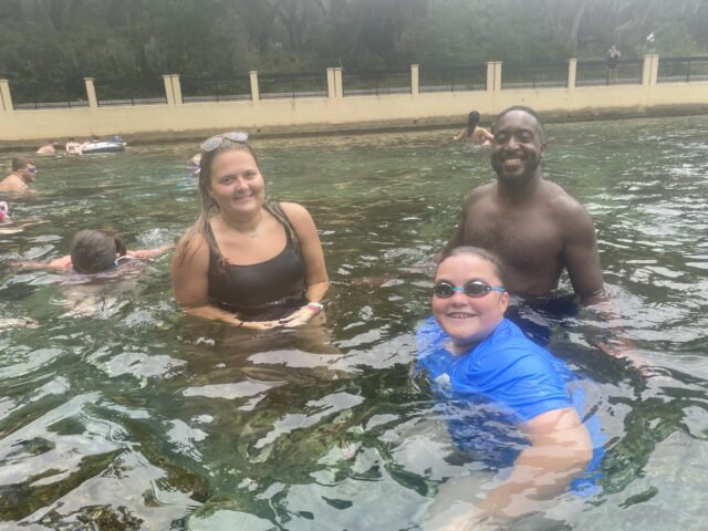 A group of people enjoying a swim in a natural water setting. They are smiling and wearing swimwear, with trees and a fence visible in the background.