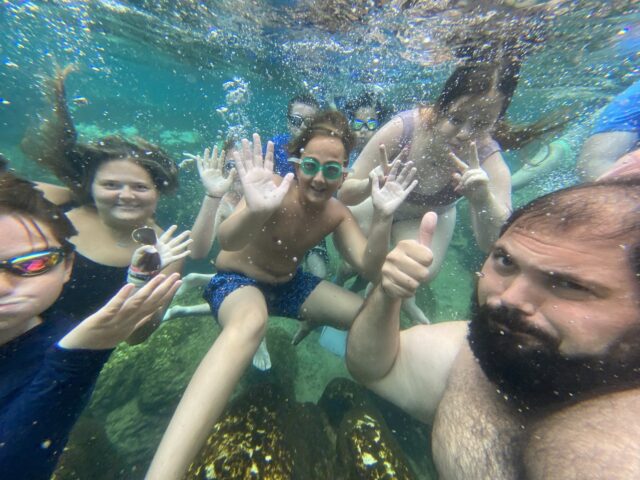 A group of people underwater in a pool, waving and giving a thumbs up, wearing swimsuits and goggles.