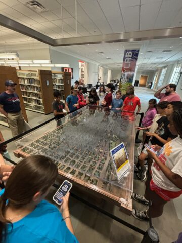 A group of people stands around a large, detailed model of a city inside a library, observing and listening to a guide. Nearby shelves are filled with books.