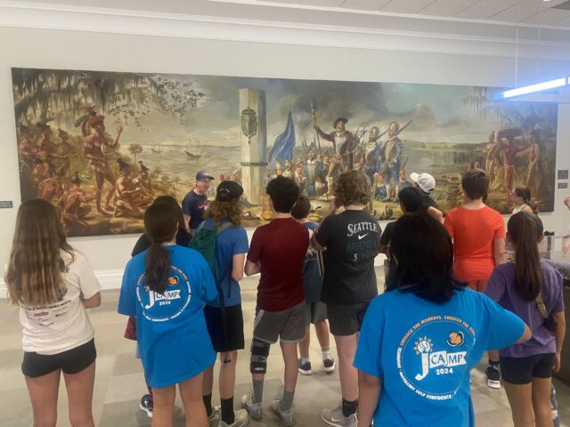 A group of people at a museum observes a large historical painting. Some are wearing colorful t-shirts with "Camp 2024" printed on them.