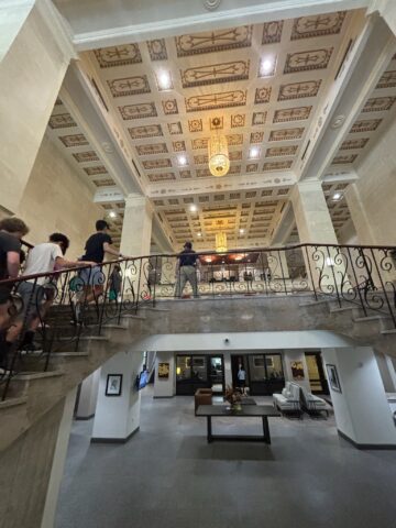 A group of people walk up a grand staircase in a lavish lobby, featuring an ornate ceiling and chandelier.