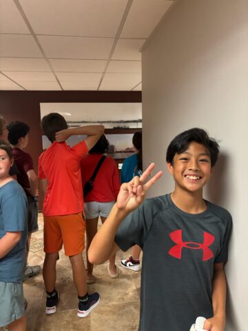 Boy in a gray shirt flashes a peace sign, smiling, with people gathered in the background.