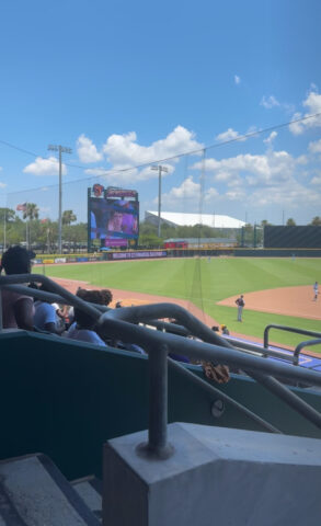 A baseball game at a stadium under a blue sky with a few clouds. Spectators are seated, watching the field. A large screen displays content in the background.