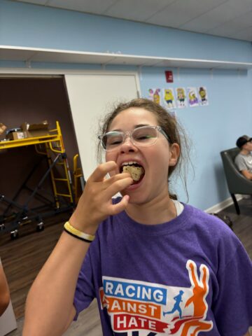 A young person wearing glasses and a purple "Racing Against Hate" shirt is eating a cookie in a room with a light blue wall.