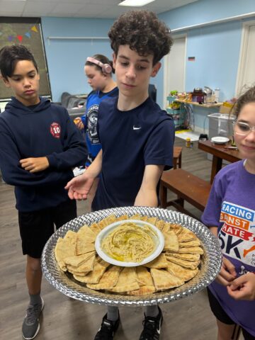 A group of four young people stand around a large platter of pita bread and dip in a room with tables and chairs.