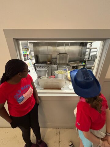 Two people wearing red shirts and blue hats look through a window into a commercial kitchen with various cooking equipment and ingredients visible.